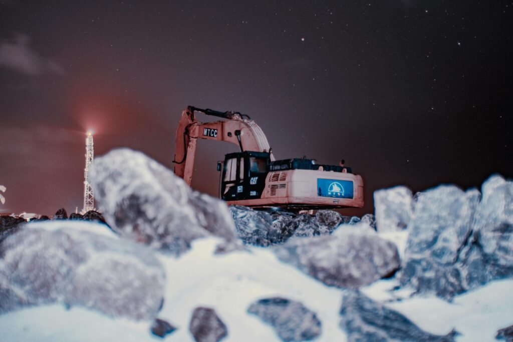 A nighttime scene featuring an excavator on snow-covered rocks under a starry sky.